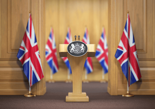 An empty wooden podium with two microphones and the UK Royal Coat of Arms, flanked by Union Jack flags in an official briefing room.